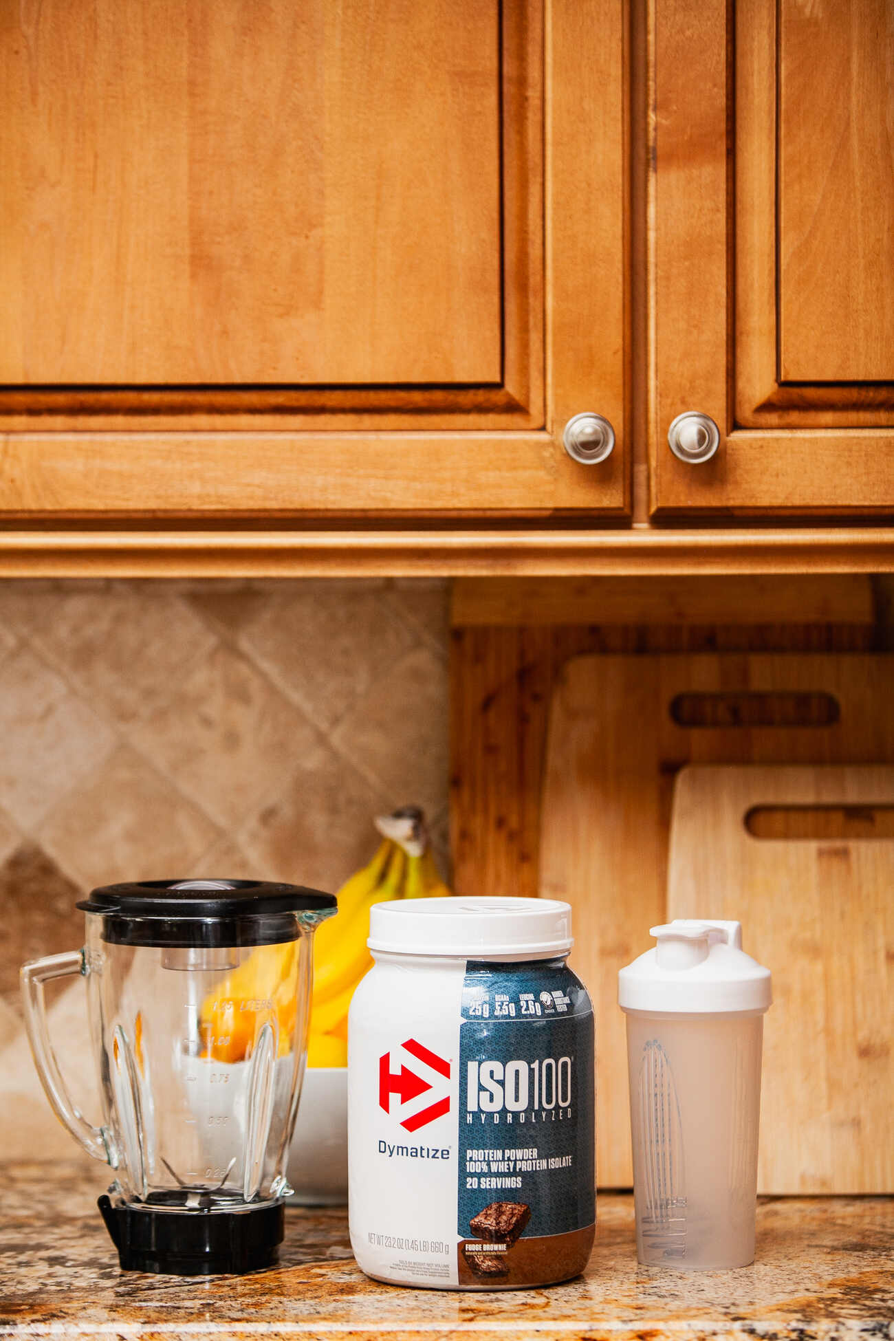 A kitchen counter with a protein powder jar, a blender, and a shaker bottle, with wooden cabinets in the background