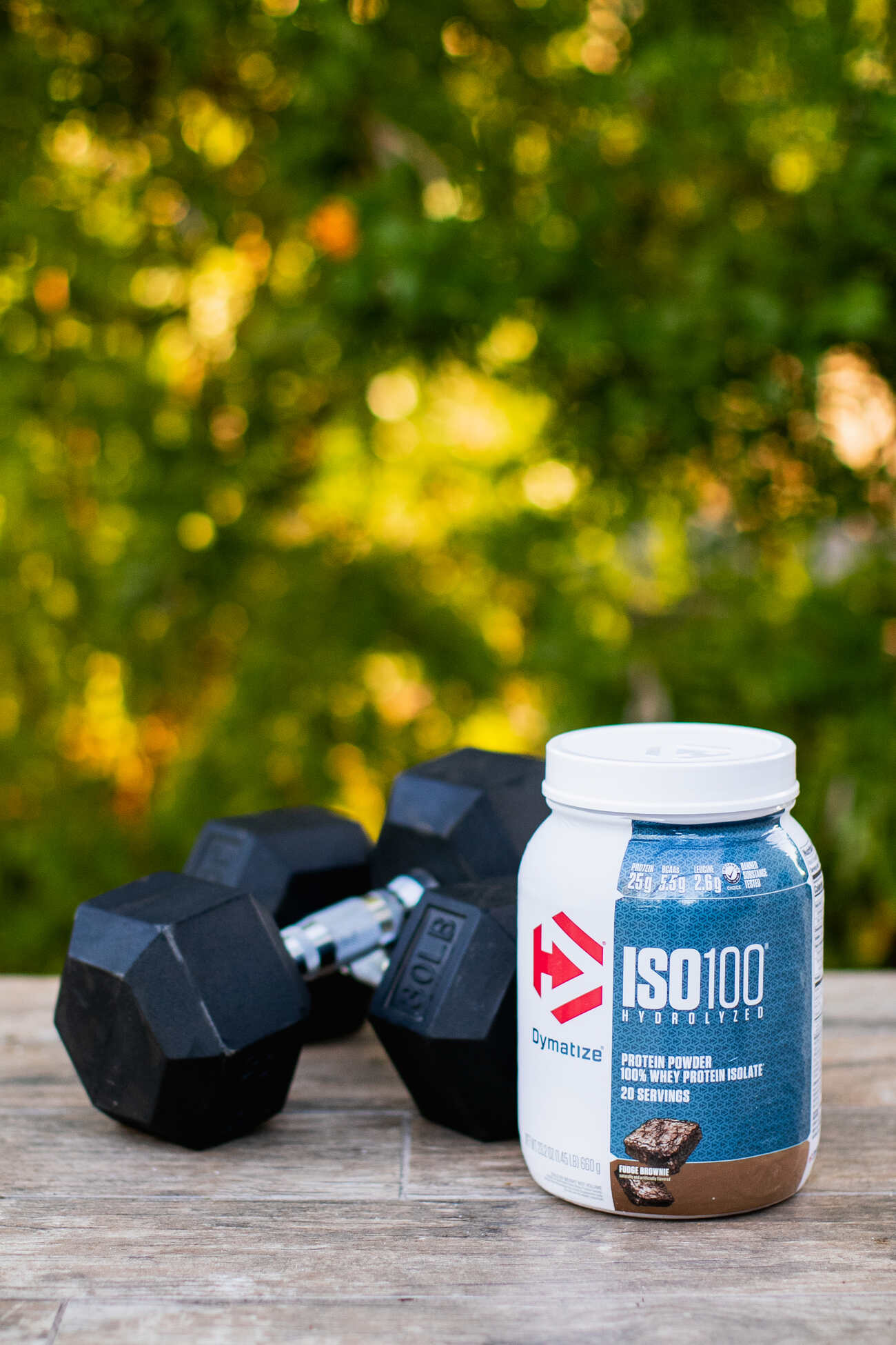Fitness equipment and a protein powder jar on a wooden table outdoors, with green foliage in the background