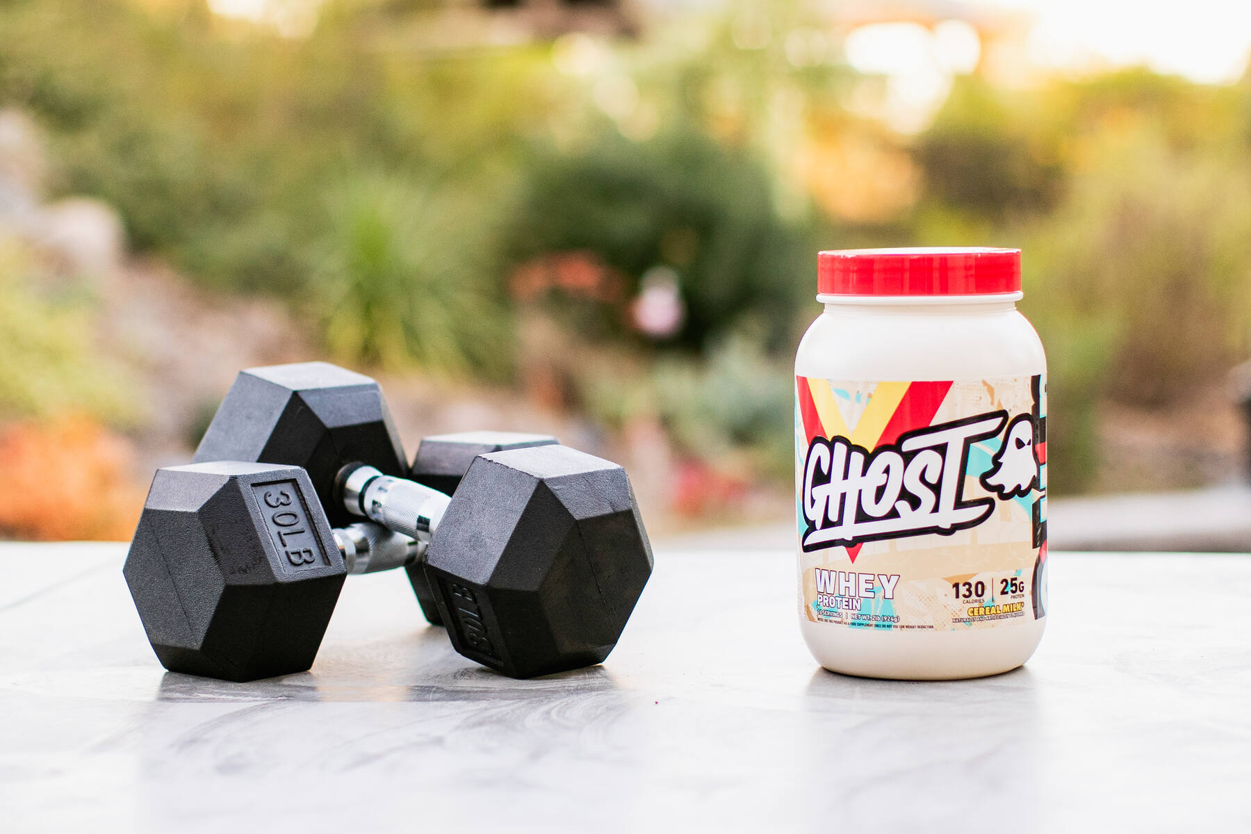 An outdoor setting with a container of Ghost Whey Protein in a vibrant, graffiti-style labeled bottle next to a pair of black hexagonal 30lb dumbbells, with greenery in the soft-focus background