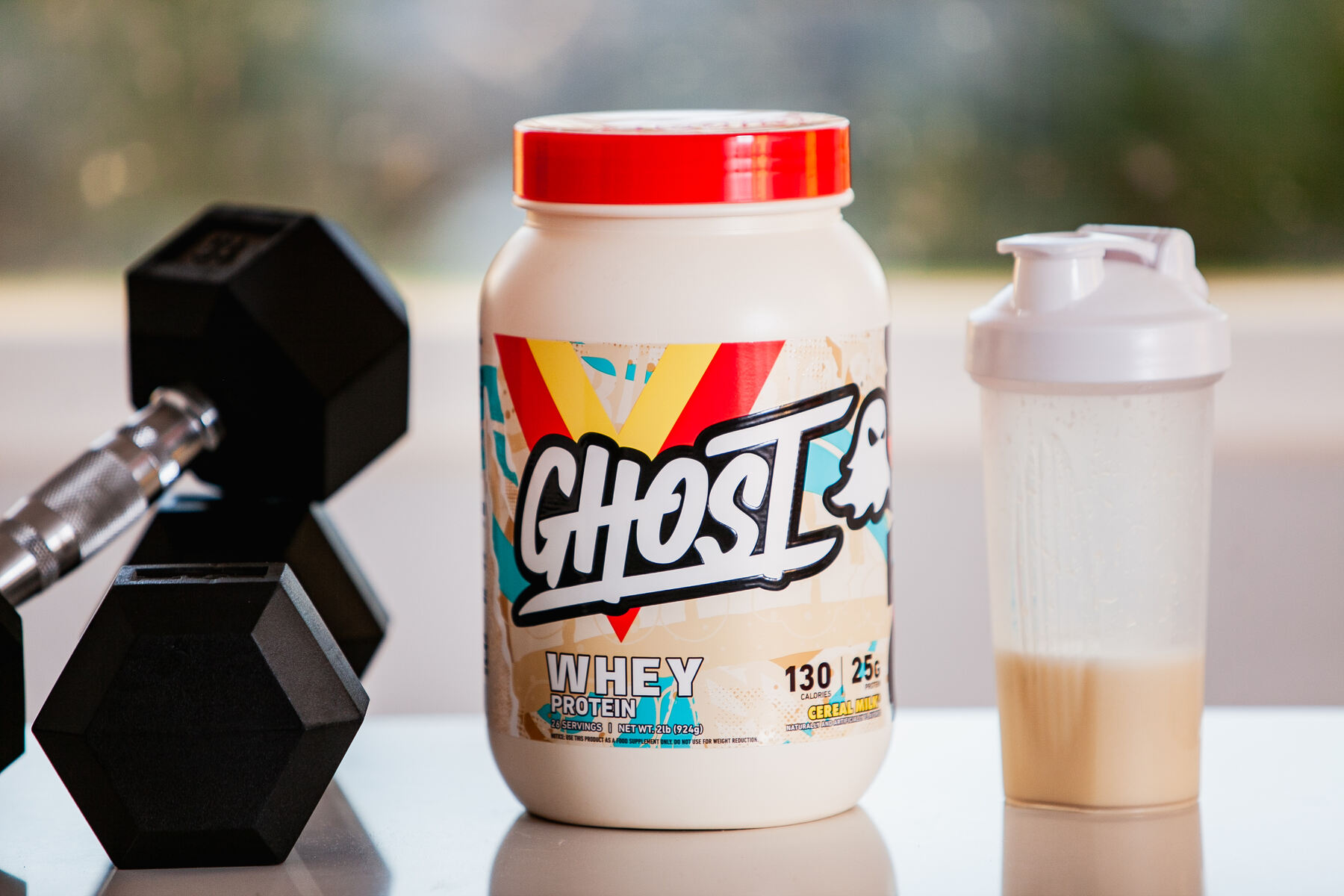 A container of Ghost Whey Protein with a colorful label next to a black dumbbell and a white protein shaker filled with a beige liquid, set against a blurred natural backdrop