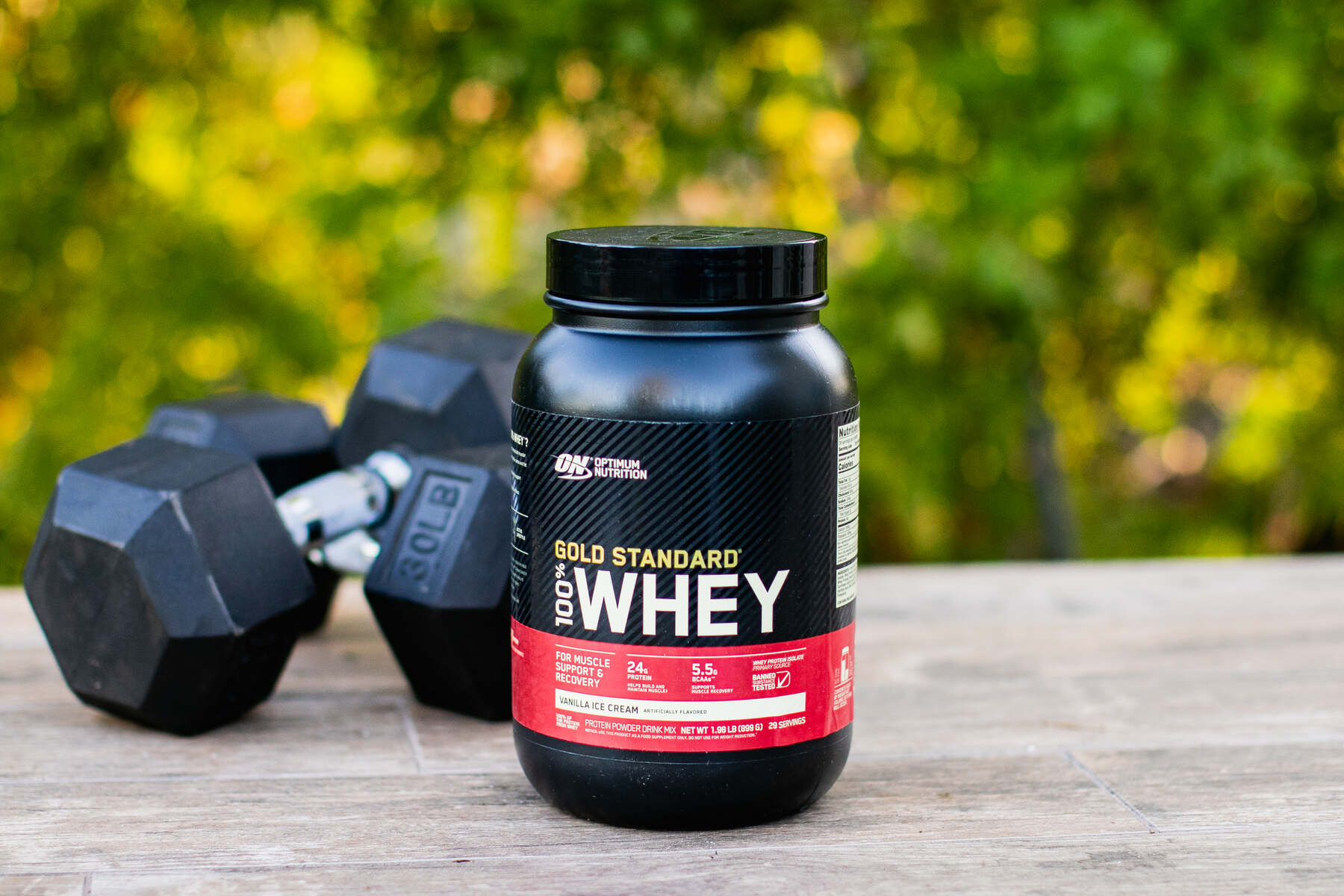 A black container of Gold Standard Whey Protein powder with a red label sits on a wooden surface next to black dumbbells with green foliage in the background