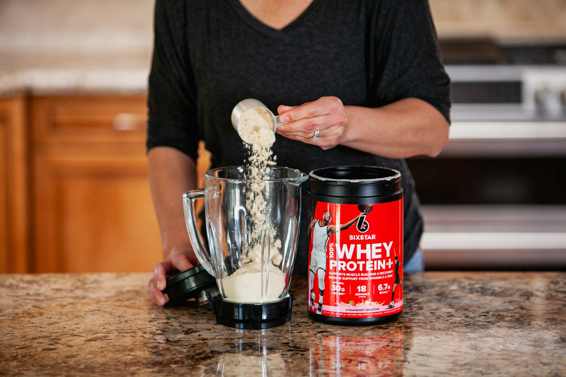 Person in a black top pouring whey protein powder into a blender next to a red container of Six Star Whey Protein on a kitchen counter