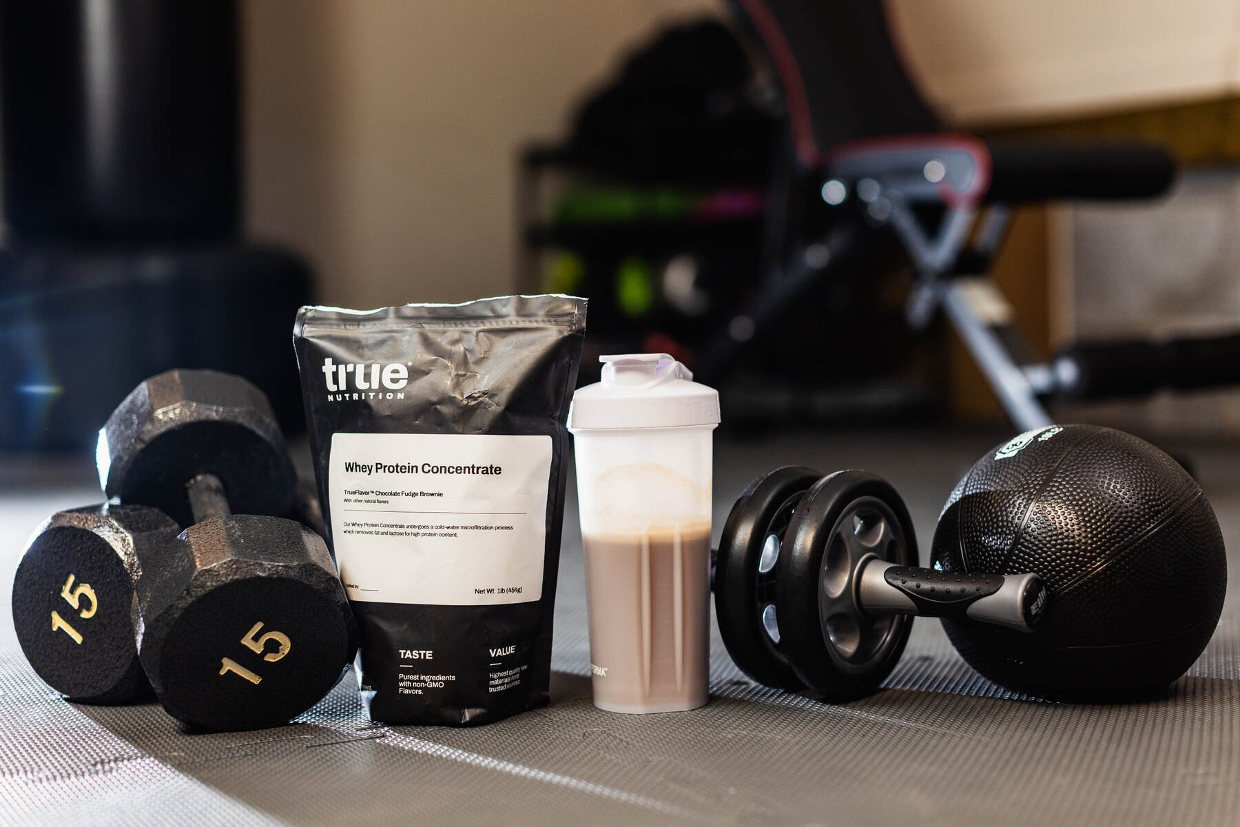 A home gym scene with dumbbells, a protein shake, and a medicine ball, alongside a bag of chocolate fudge brownie whey protein