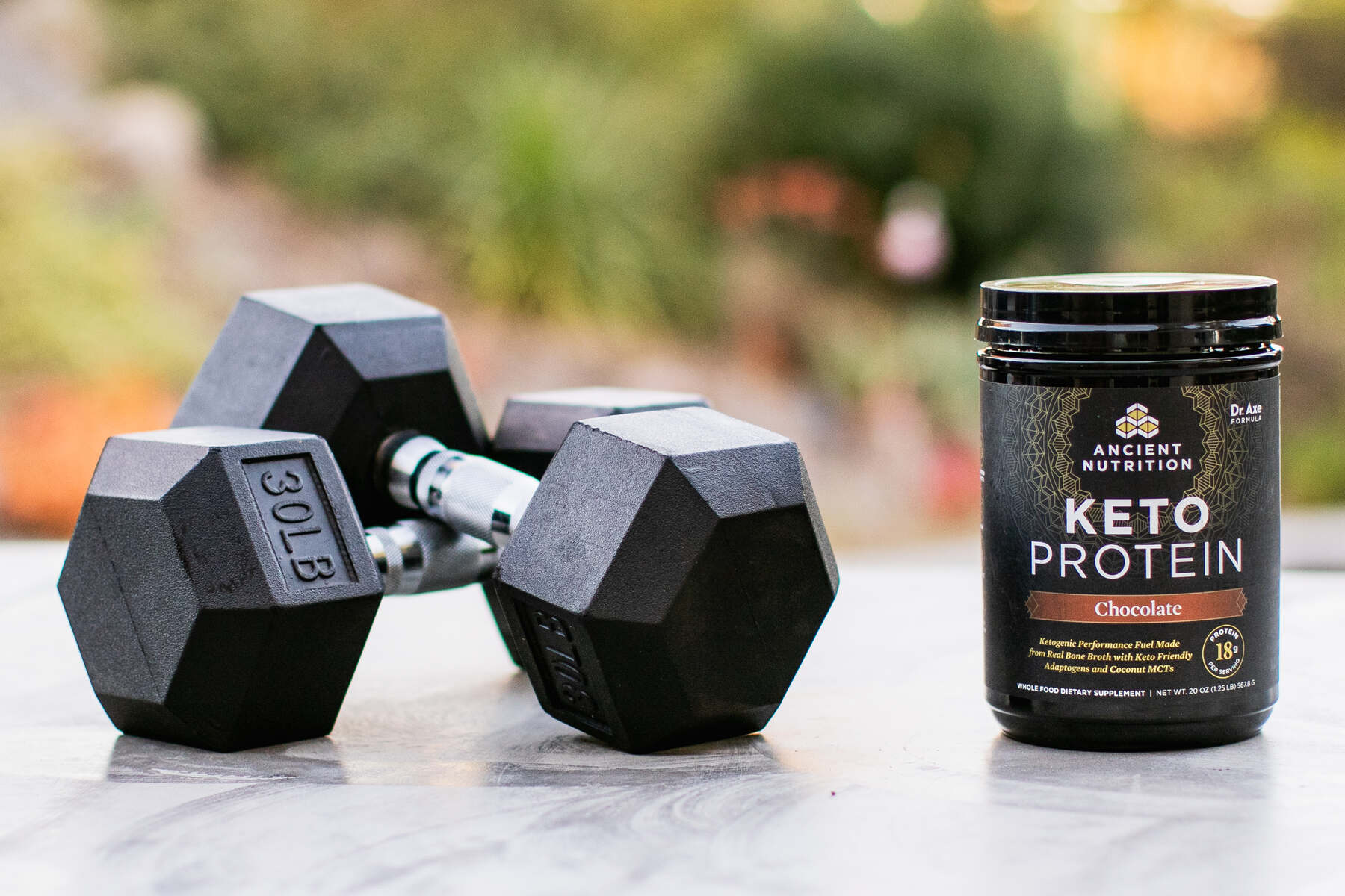 Outdoor setting with a container of Ancient Nutrition Keto Protein in chocolate flavor next to a pair of 30lb dumbbells