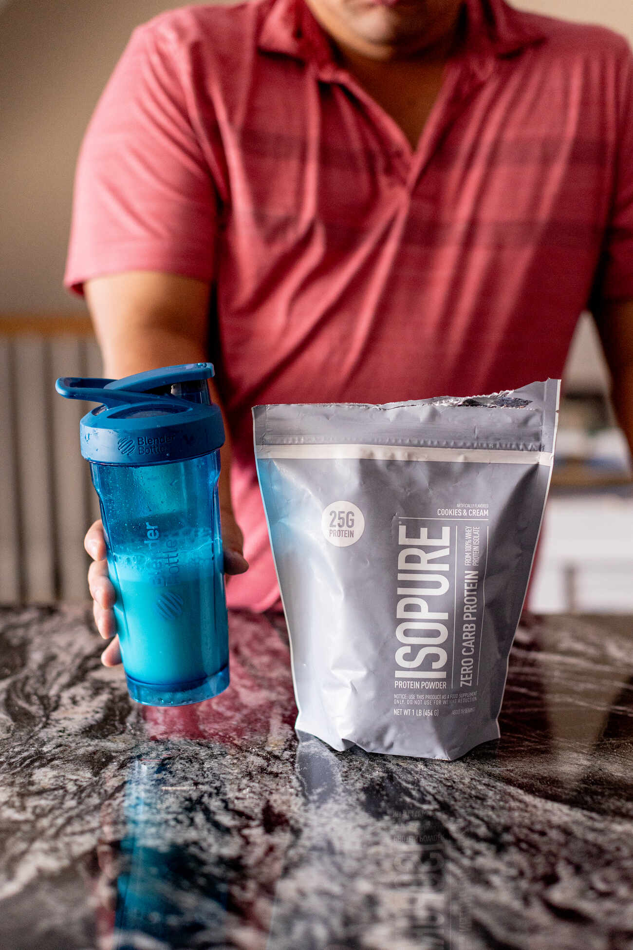 Main holding a protein shake in a blue Bottle Blender next to a bag of Isopure protein powder on a black and white granite countertop