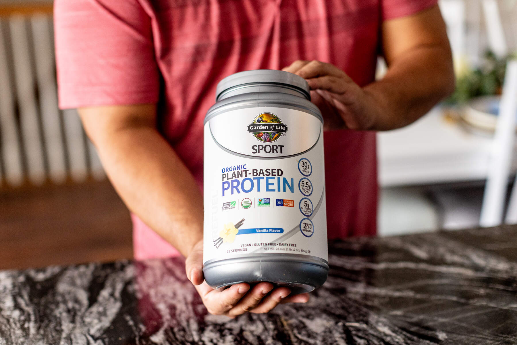 A man holding a jar of organic plant-based protein powder in a kitchen setting