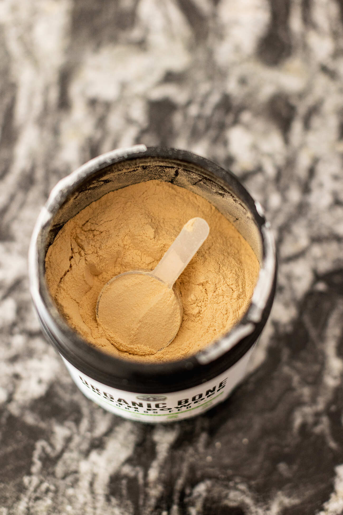 A top view of an open jar of bone broth protein powder with a scoop on a marbled surface
