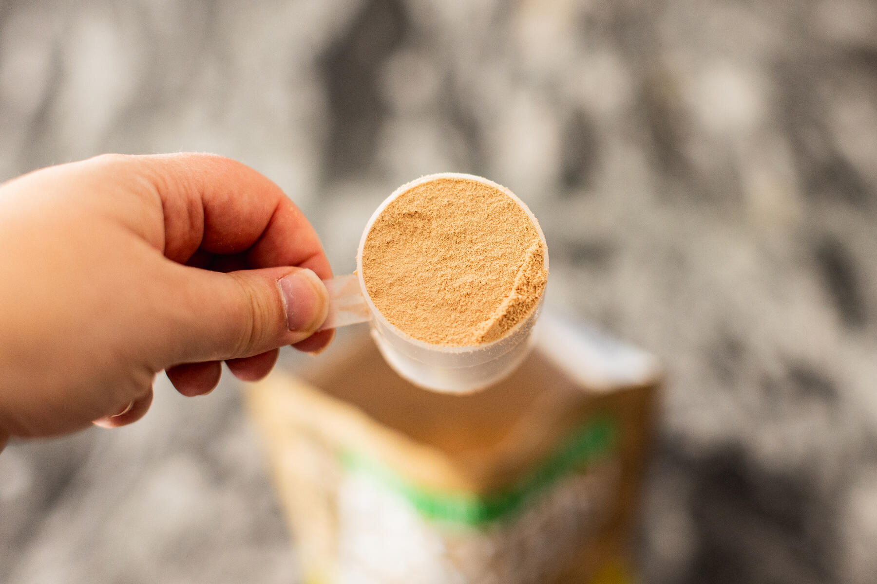 Person holding a full scoop of protein powder above a granite countertop