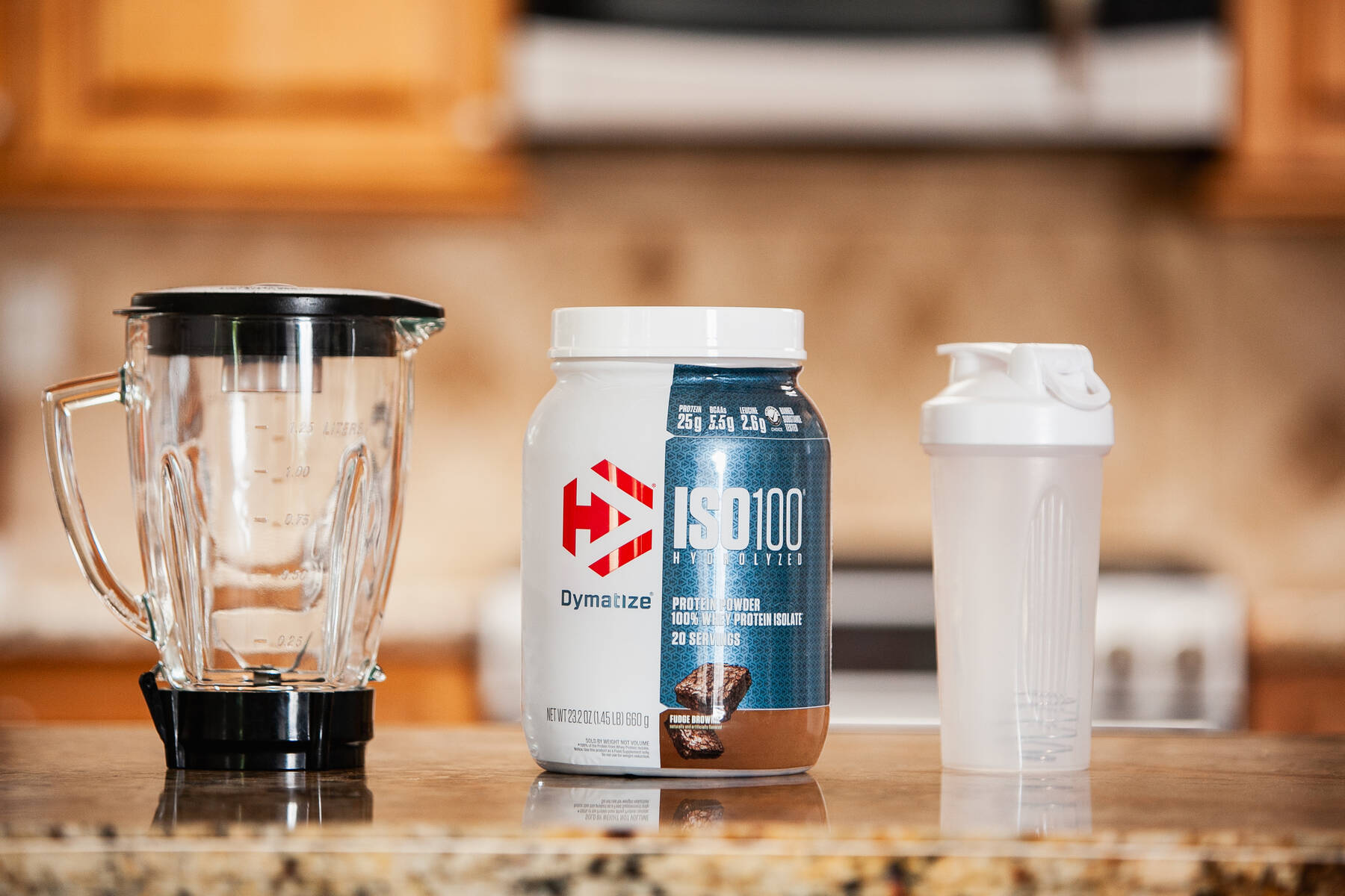 A kitchen countertop displaying a clear blender, a white and blue container of Dymatize ISO100 Hydrolyzed protein powder, and a white protein shaker