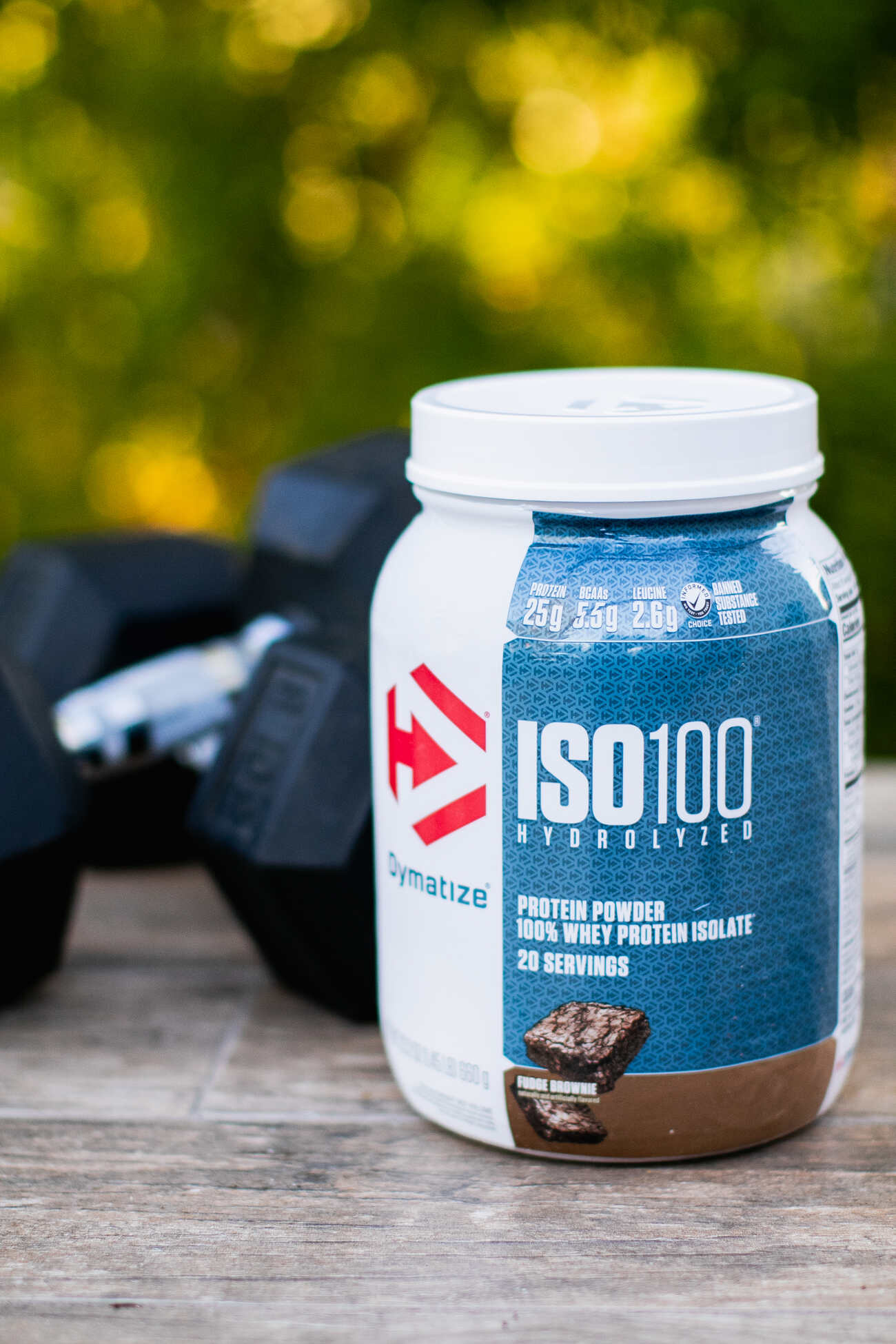 A white and blue container of Dymatize ISO100 Hydrolyzed protein powder, placed on an outdoor surface with a pair of black dumbbells to the side and green foliage in the background