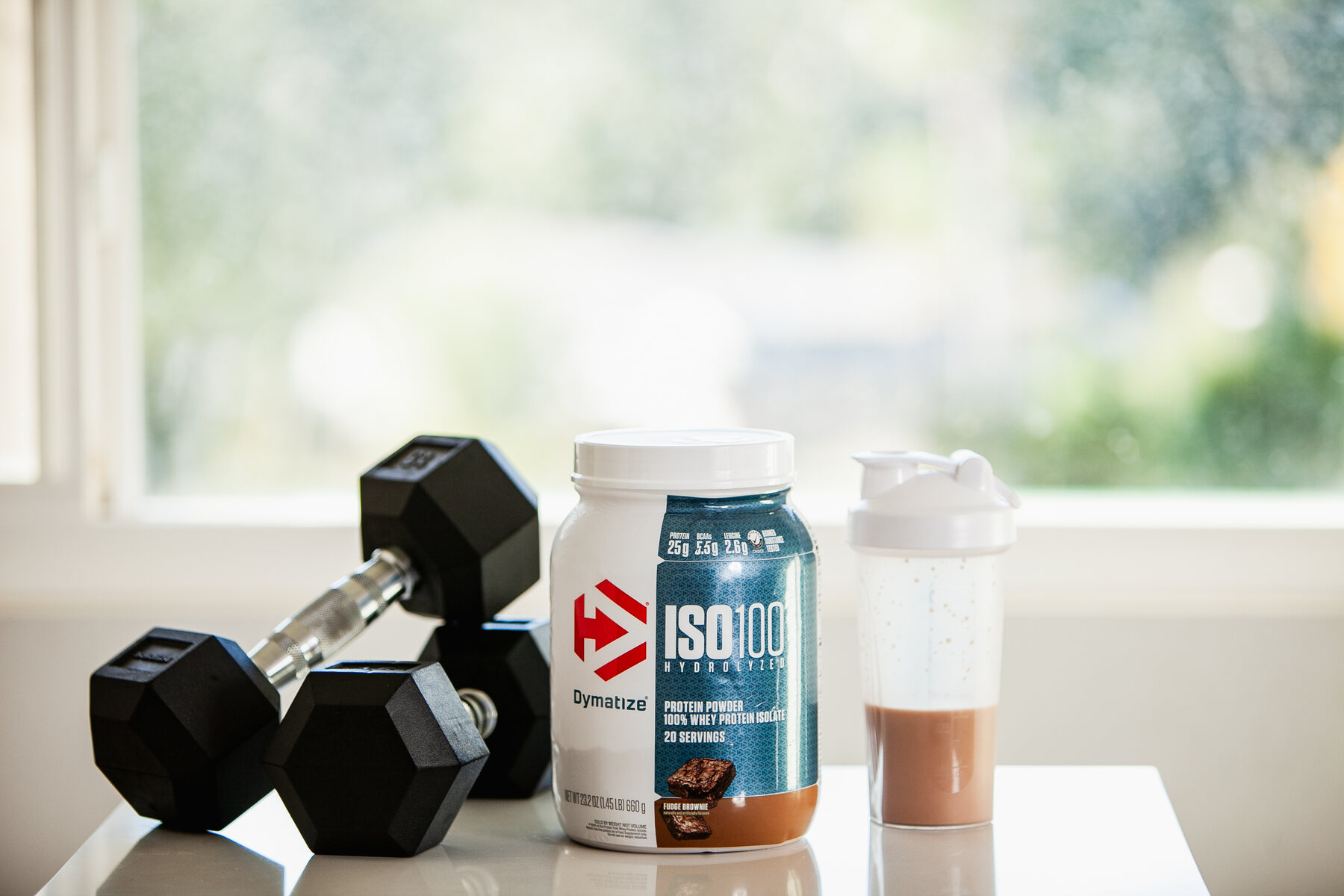 A black dumbbell, a jar of protein powder, and a filled shaker bottle on a white surface with a window in the background