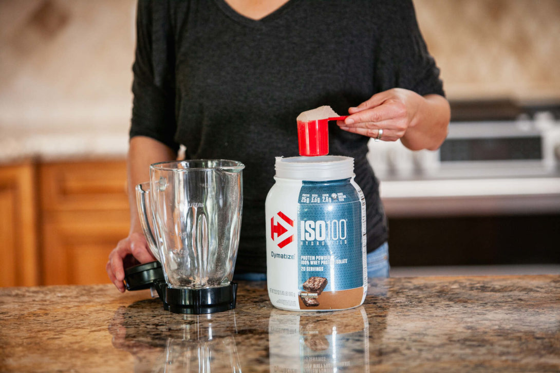 A woman scooping protein powder from a jar into a blender on a kitchen counter