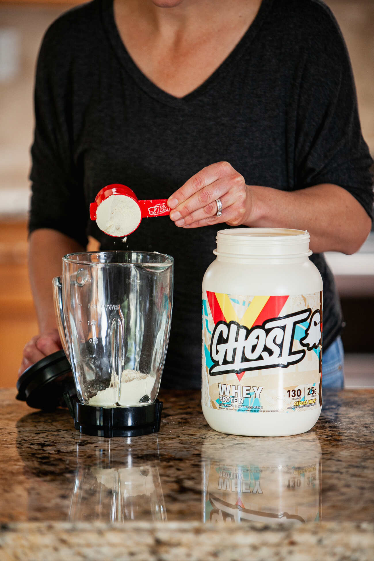 A person in a black top is scooping powder from a Ghost Whey Protein container into a blender on a kitchen countertop