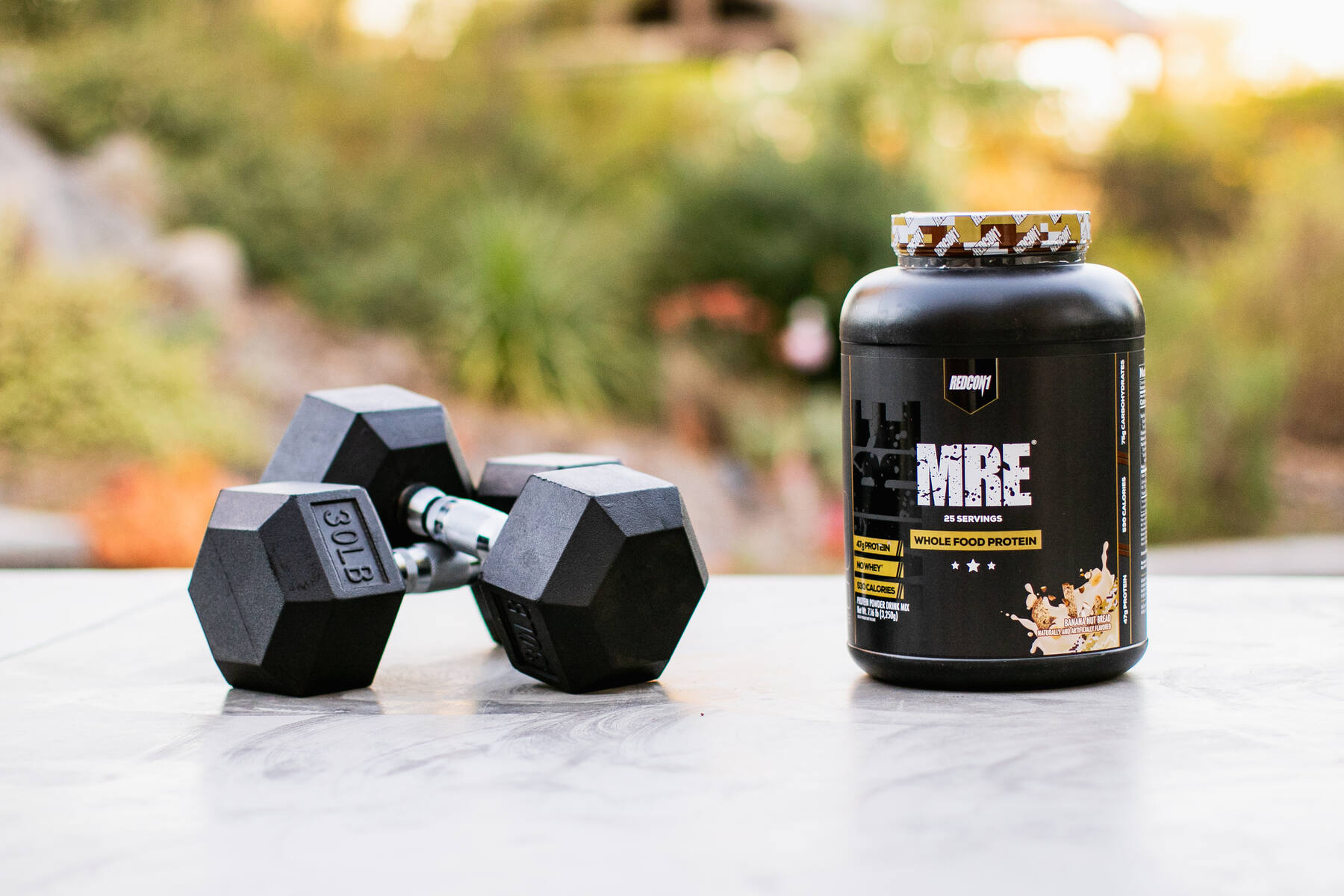 An outdoor setting with a black container of Redcon1 MRE Whole Food Protein on a surface next to a pair of black dumbbells, with natural greenery in the background