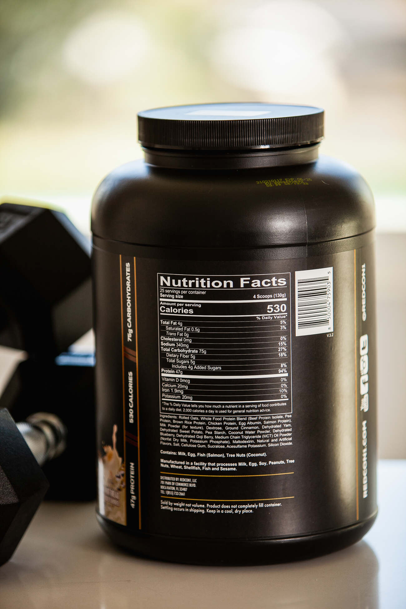 A close-up of the nutritional label on the back of a black container of Redcon1 MRE Whole Food Protein, detailing the calorie content and ingredients, with a dumbbell in the blurred background