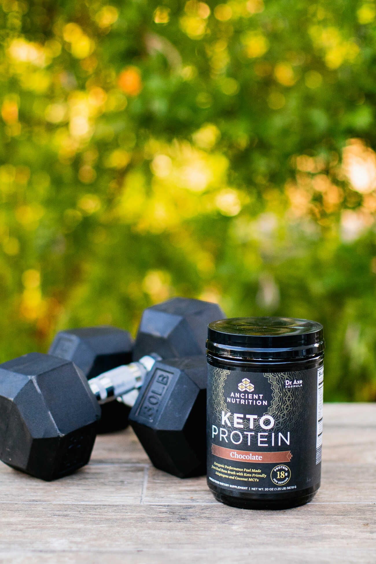 A black dumbbell and a jar of keto protein powder on a wooden surface with greenery in the background