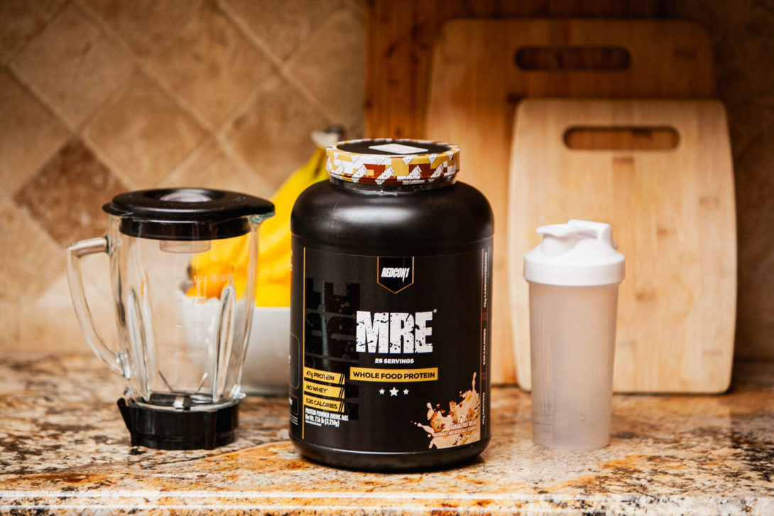 A kitchen counter with a jar of protein powder, a blender, and a shaker bottle, with wooden cabinets in the background
