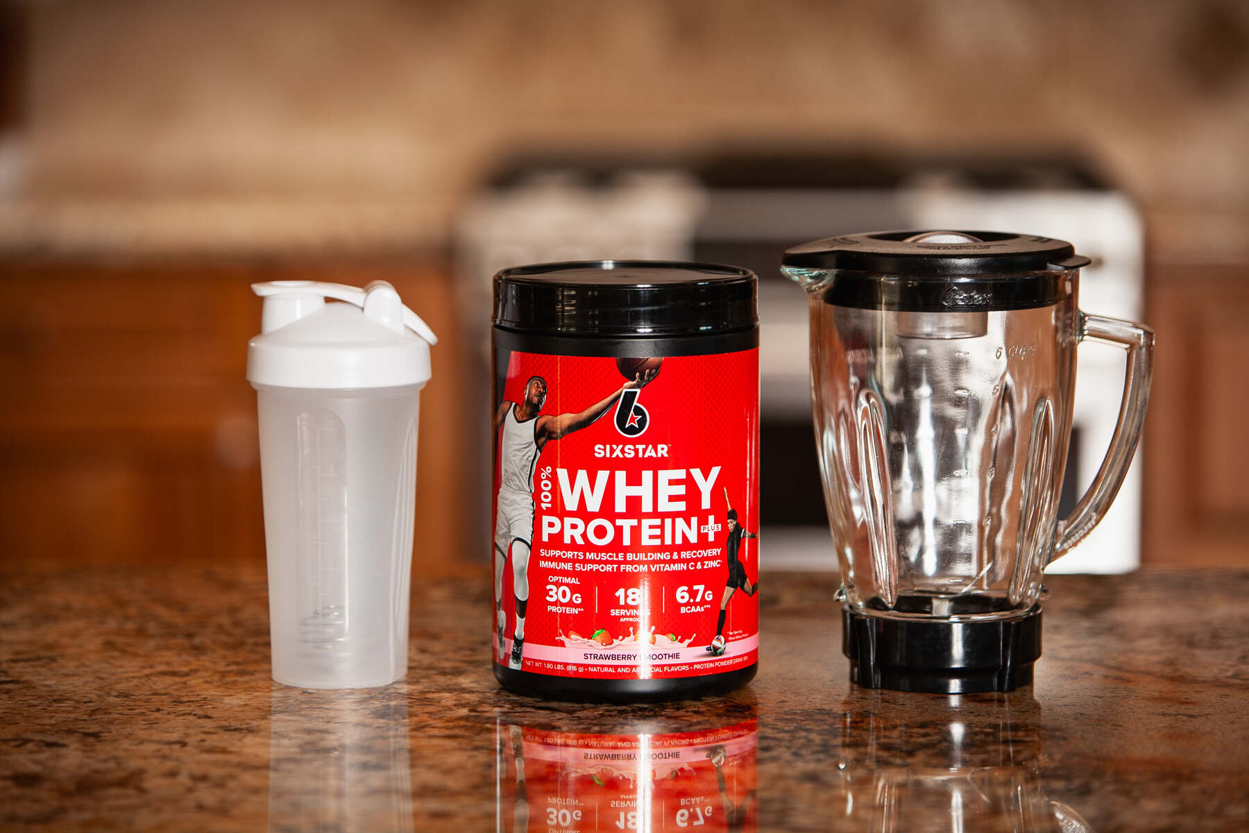 A protein powder jar, shaker bottle, and blender on a kitchen counter with wooden cabinets