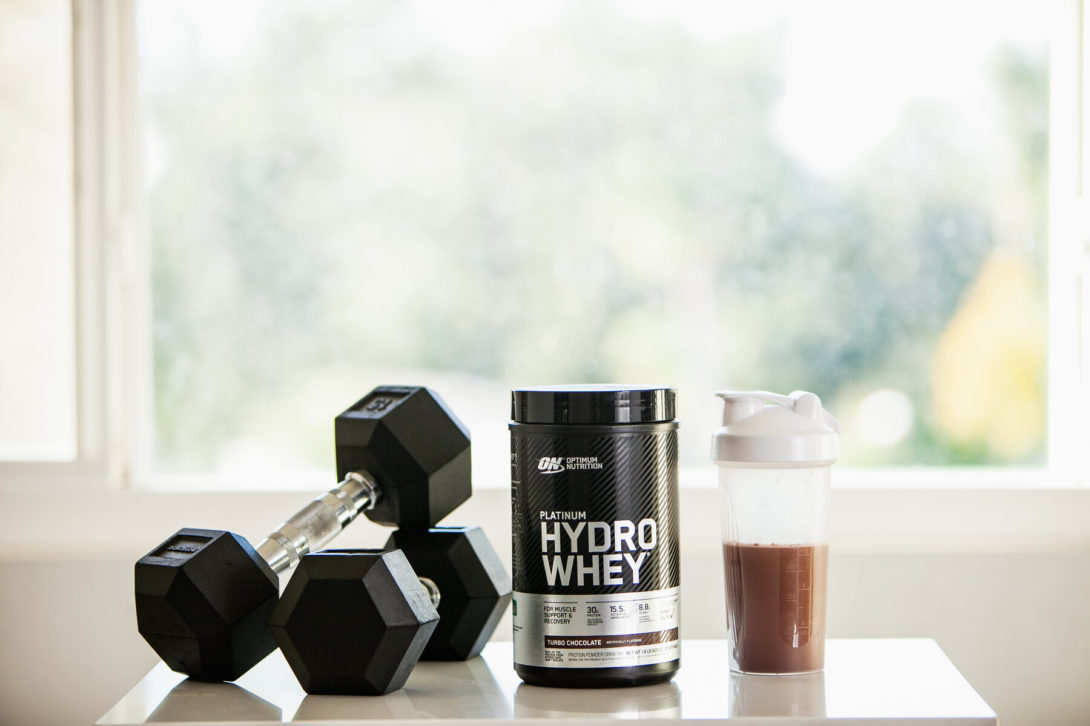 A black container of Optimum Nutrition Platinum Hydro Whey protein powder next to a black dumbbell and a white protein shaker filled with a chocolate-flavored drink, set against a bright window backdrop