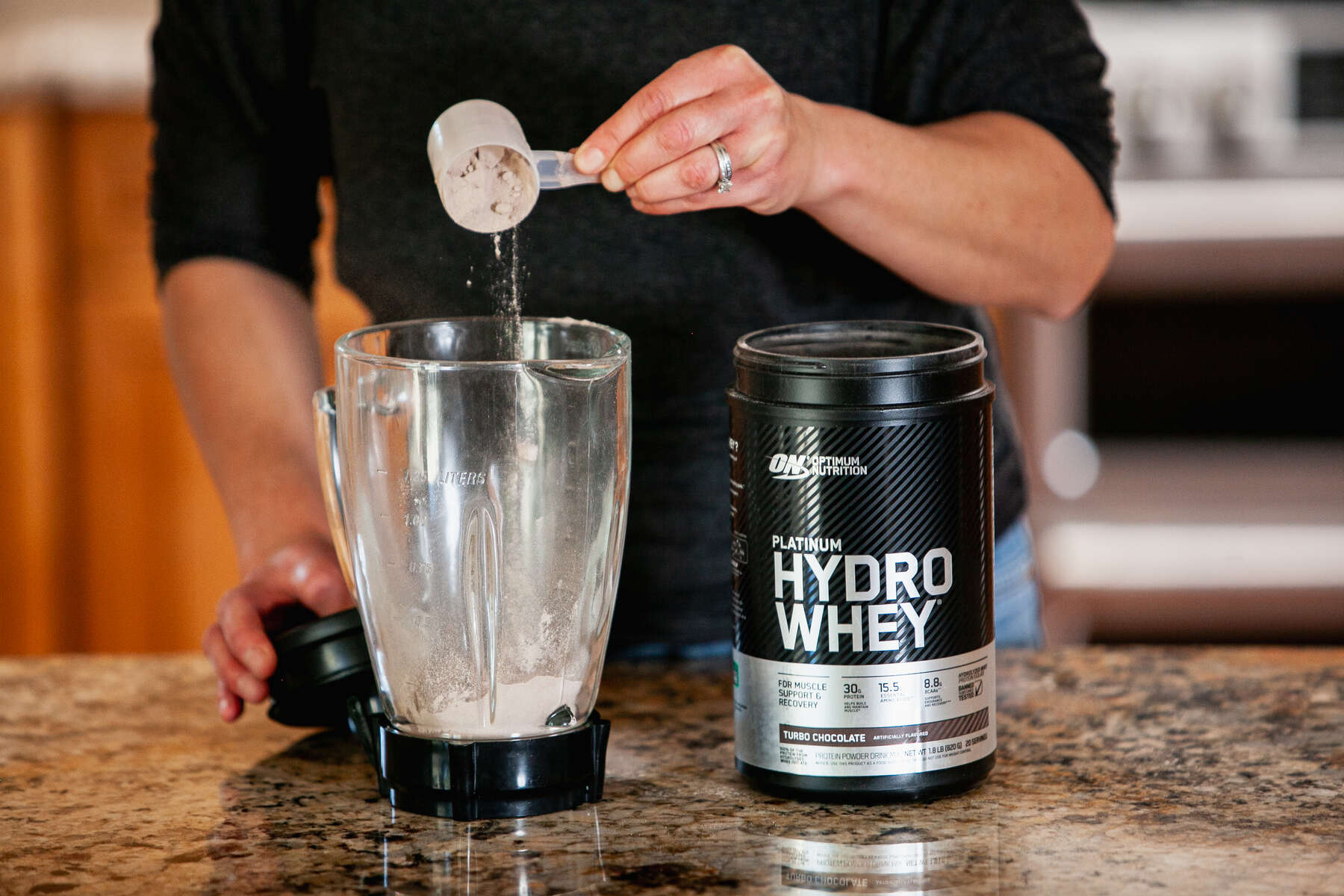 A person in a black top is pouring protein powder from a measuring scoop into a blender next to a black container of Optimum Nutrition Platinum Hydro Whey on a kitchen counter.