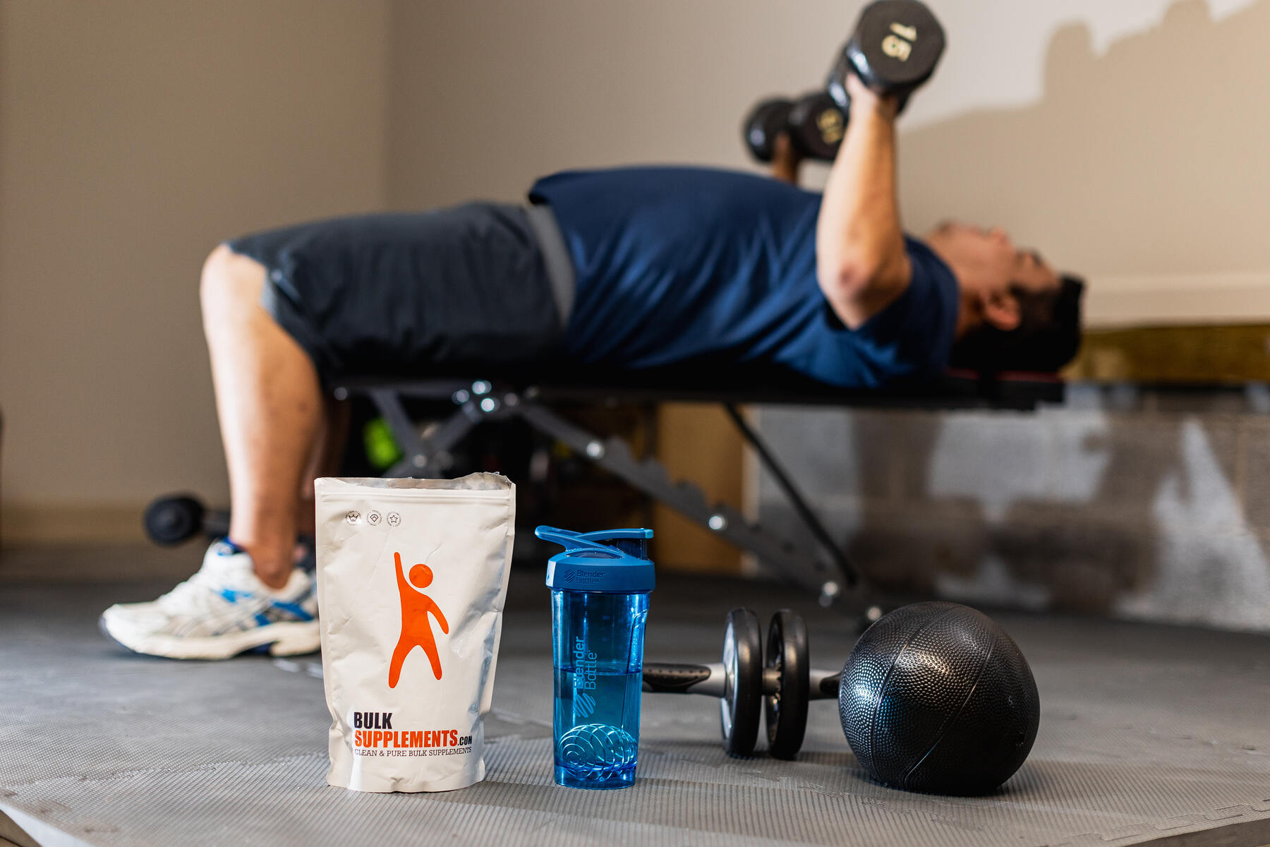 Bulk Supplements bag next to a water bottle and fitness equipment with a man lifting weights in the background