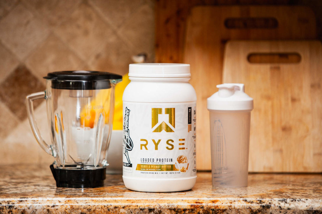 A protein powder jar, blender, and shaker bottle on a kitchen countertop