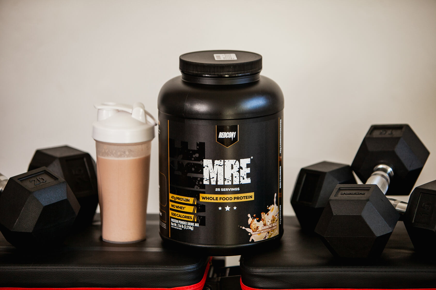 A black container of Redcon1 MRE Whole Food Protein on a workout bench next to a white protein shaker and a pair of black dumbbells, set against a neutral background