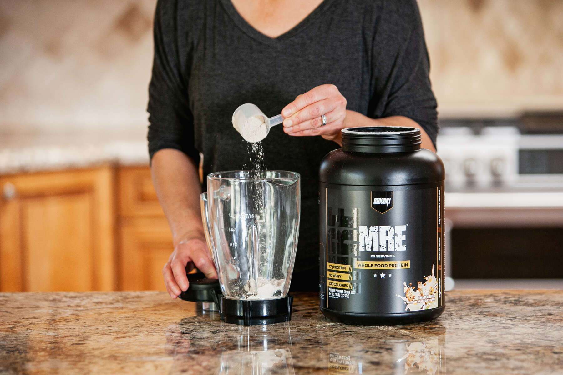 A person in a black top is scooping powder from a black container of Redcon1 MRE Whole Food Protein into a blender on a kitchen counter