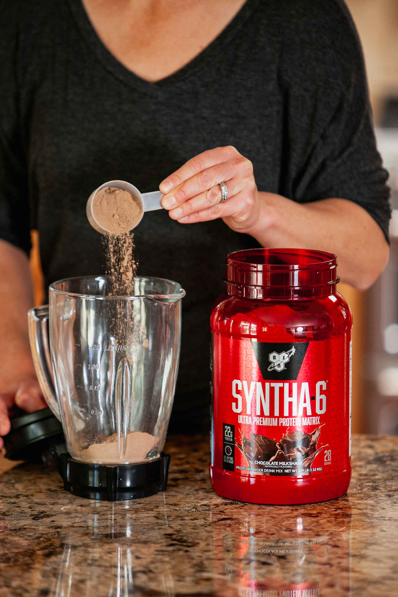A person wearing a black top is pouring chocolate-colored Syntha-6 protein powder into a blender, with a red container of the protein powder visible on a kitchen counter