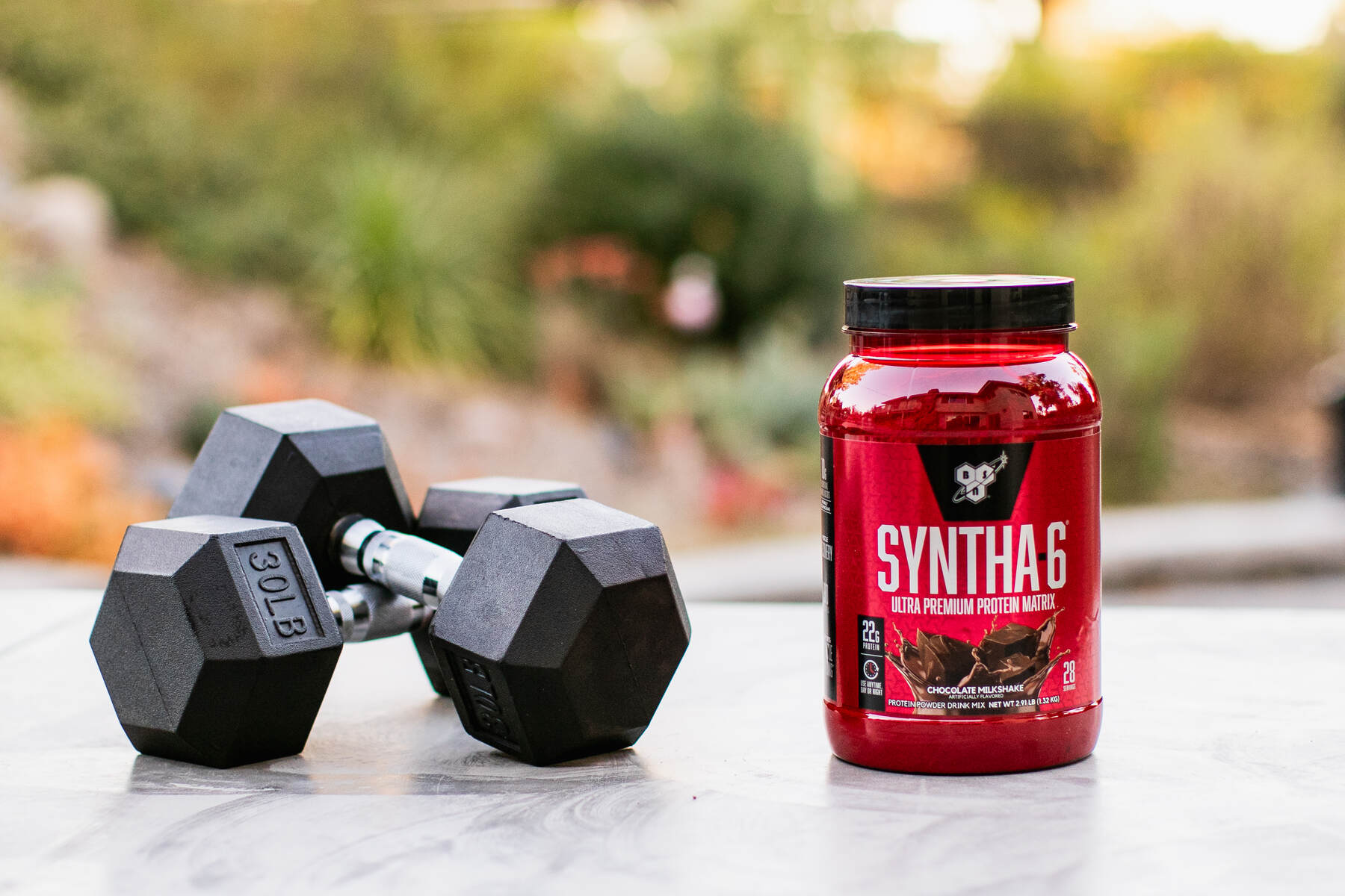 A black dumbbell and a red jar of protein powder on an outdoor marble surface with plants in the background