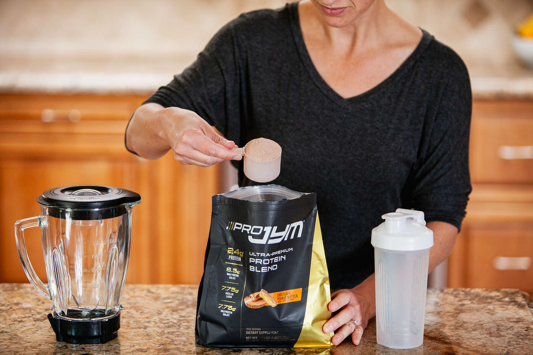 A woman scooping protein powder over a blender next to a shaker bottle on a kitchen counter