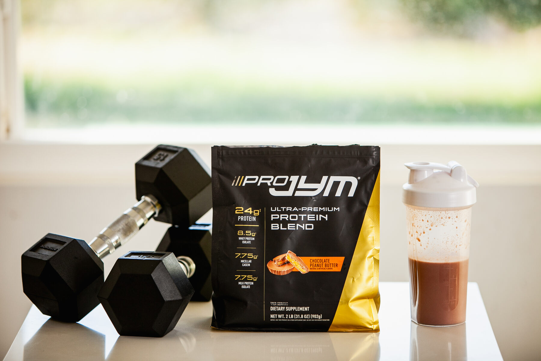 Pro Jym Protein supplement bag and shaker next to a dumbbell on a white surface