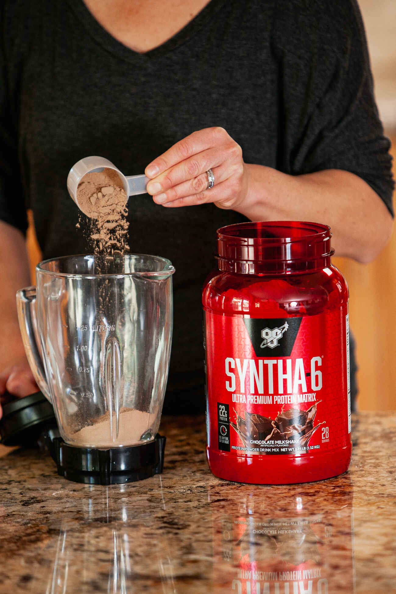 Woman in a black shirt pouring Syntha-6 protein powder into a blender on a kitchen countertop
