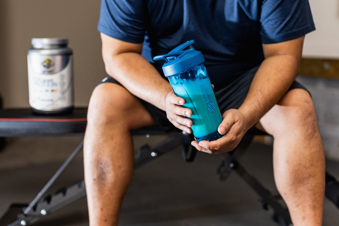 Man holding a protein shake in a blue Bottle Blender while sitting on a workout bench in a gym