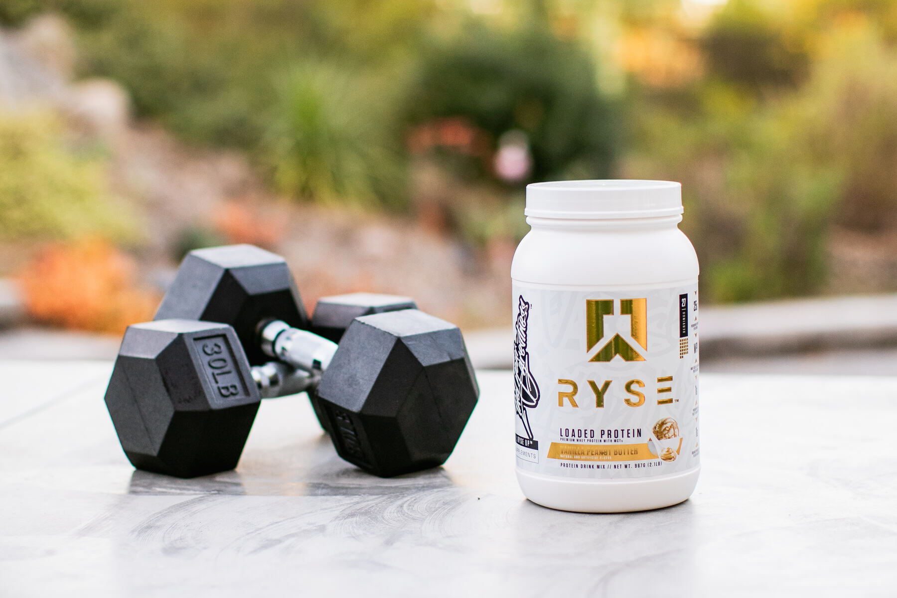 An outdoor setting with a container of RYSE Loaded Protein on a surface next to a pair of black dumbbells with green foliage in the background