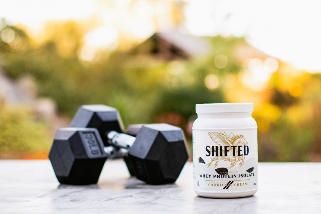 SHIFTED Whey Protein Isolate beside a pair of dumbbells on an outdoor table