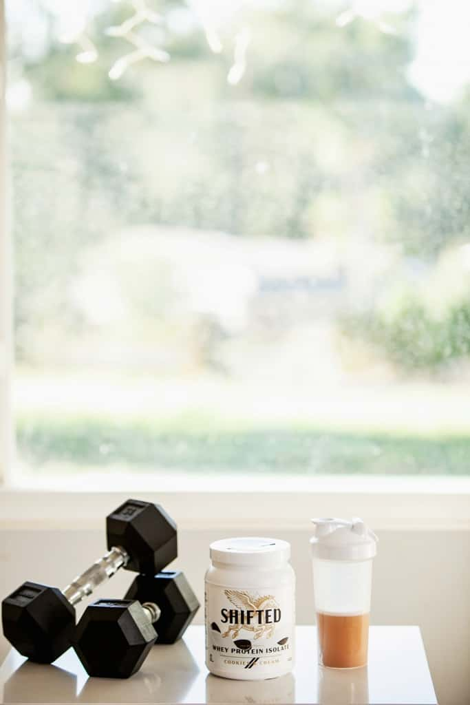 SHIFTED Whey Protein Isolate beside a pair of dumbbells and a tumbler filled with protein shake