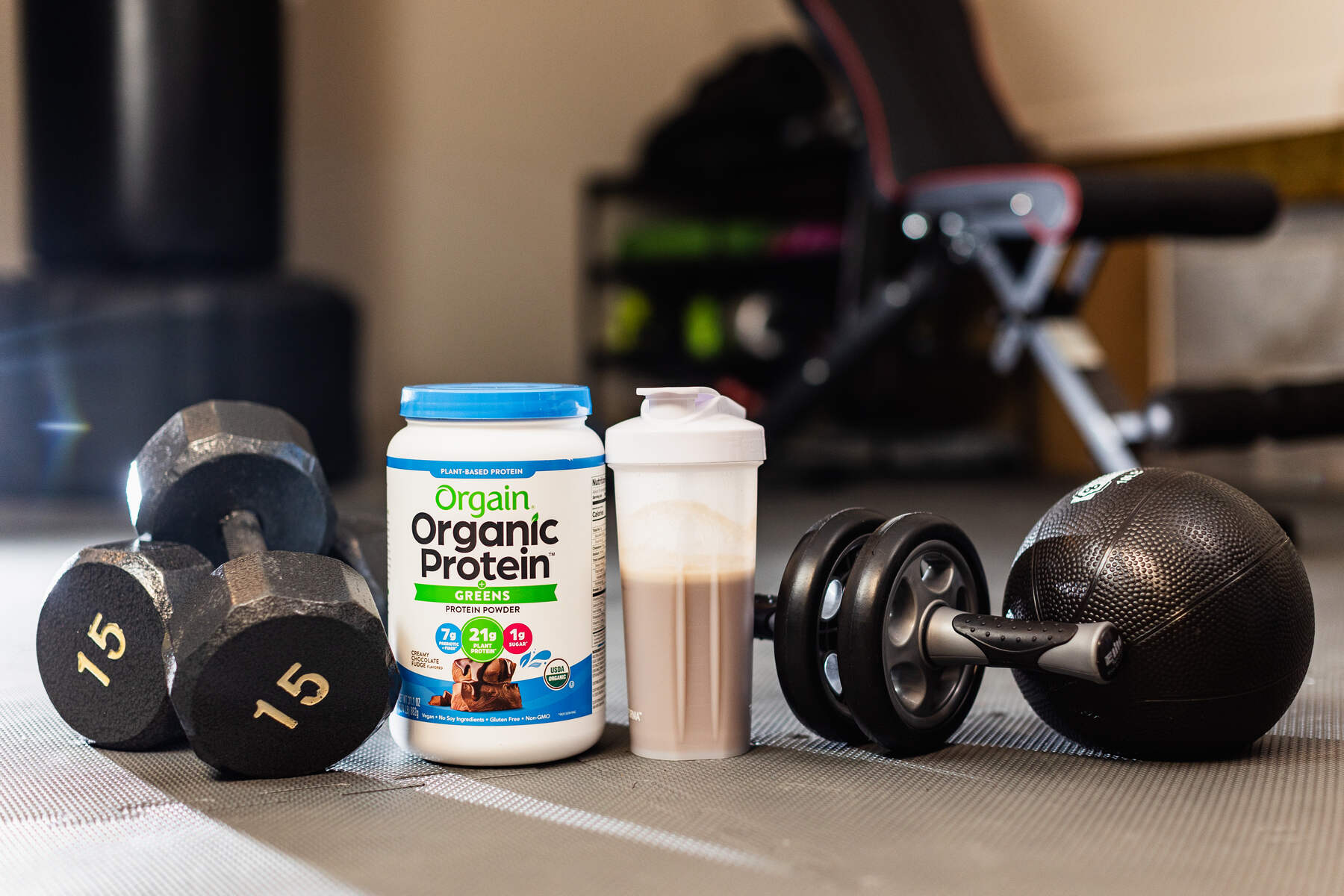 Dumbbells, a jar of organic protein powder, a shaker bottle, and exercise equipment on a gym floor