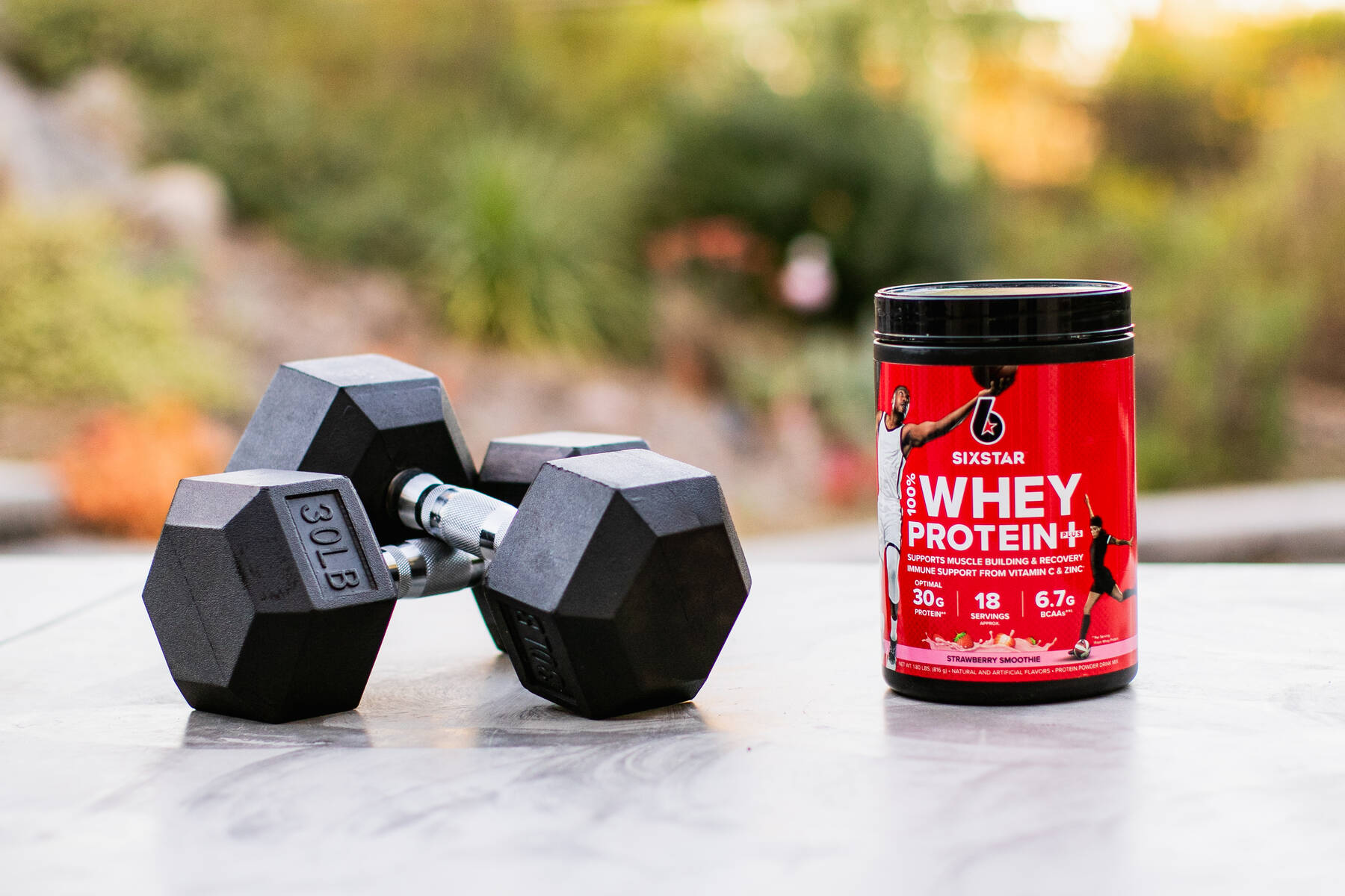 A red container of Six Star Whey Protein powder sits on an outdoor surface with a pair of black dumbbells surrounded by greenery