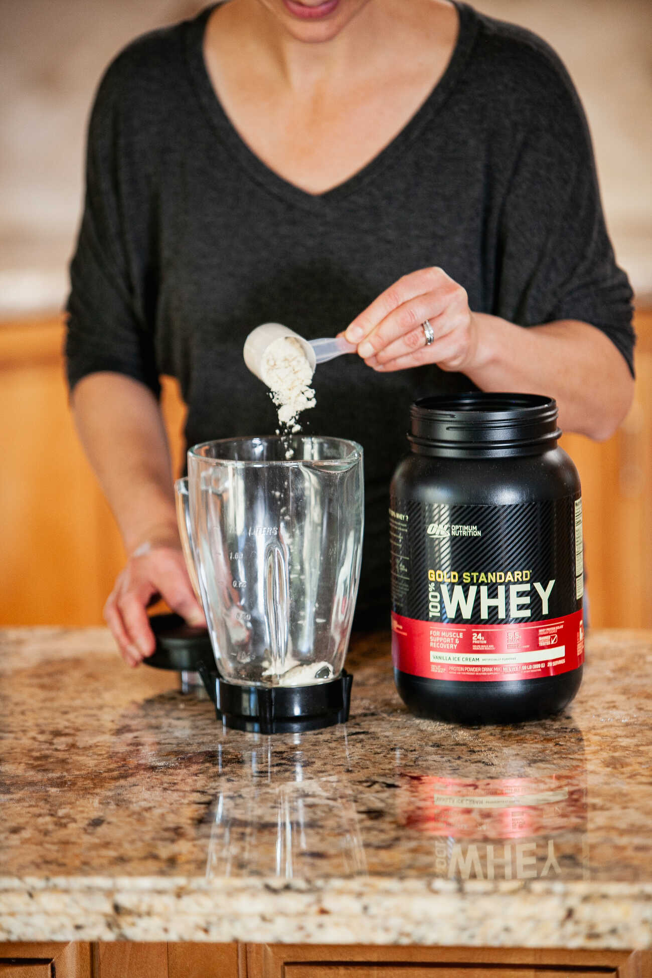 Woman in a black top scooping vanilla ice cream flavored Gold Standard Whey Protein powder into a blender on a kitchen counter