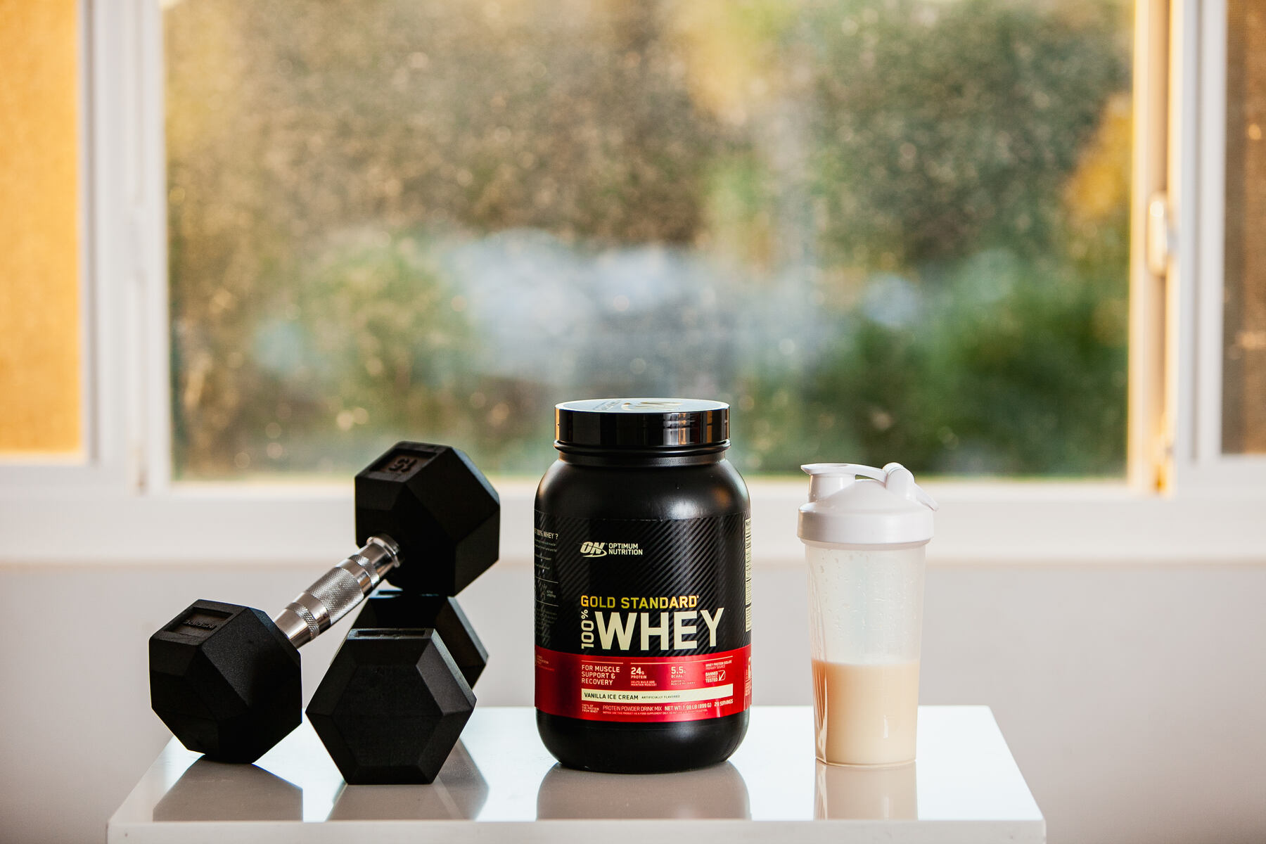 A black Gold Standard Whey Protein container and a white protein shaker filled on a white table, with a black dumbbell beside them and a window showing greenery outside.