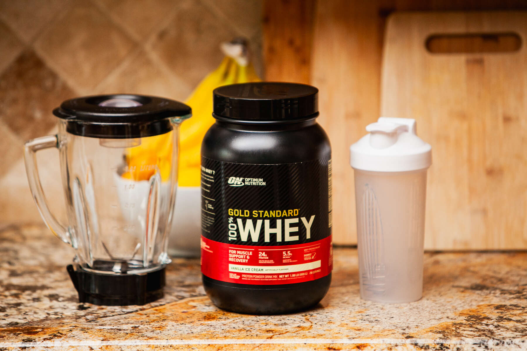 A clear blender, a black and red labeled Gold Standard Whey Protein container, and a white protein shaker on a granite countertop