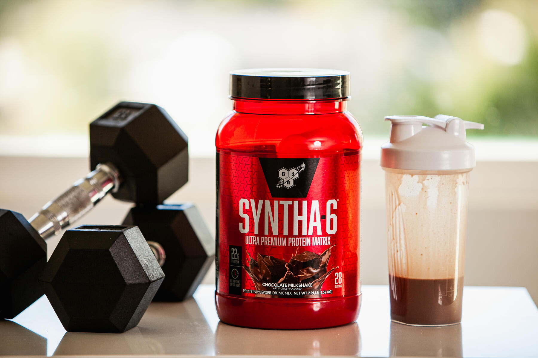 A bright red container of Syntha-6 protein powder on a white surface next to black dumbbells and a white shaker bottle with chocolate-flavored drink, with a blurred natural backdrop visible through a window