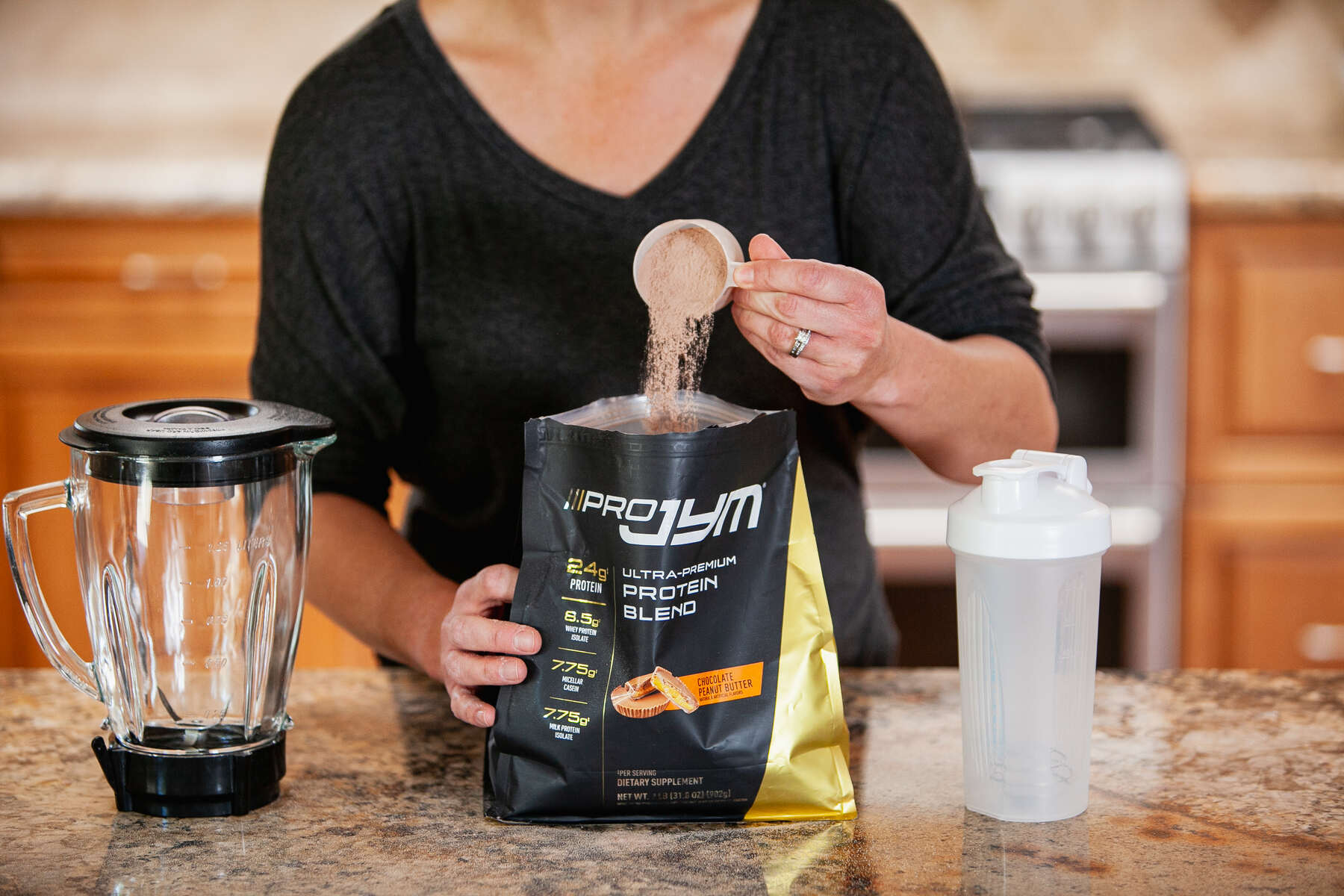 Woman in a black shirt pouring ProJym protein powder into the bag next to a blender and water bottle on a kitchen counter