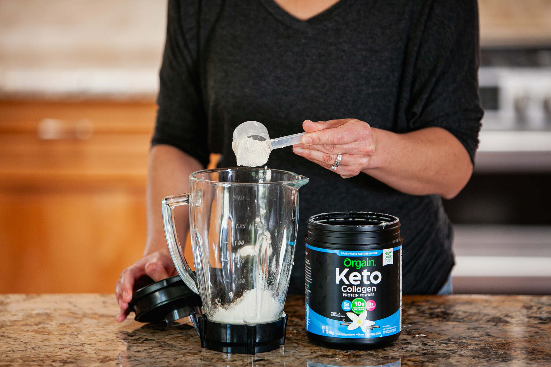 Woman pouring a scoop of vanilla protein powder into a glass blender in the kitchen