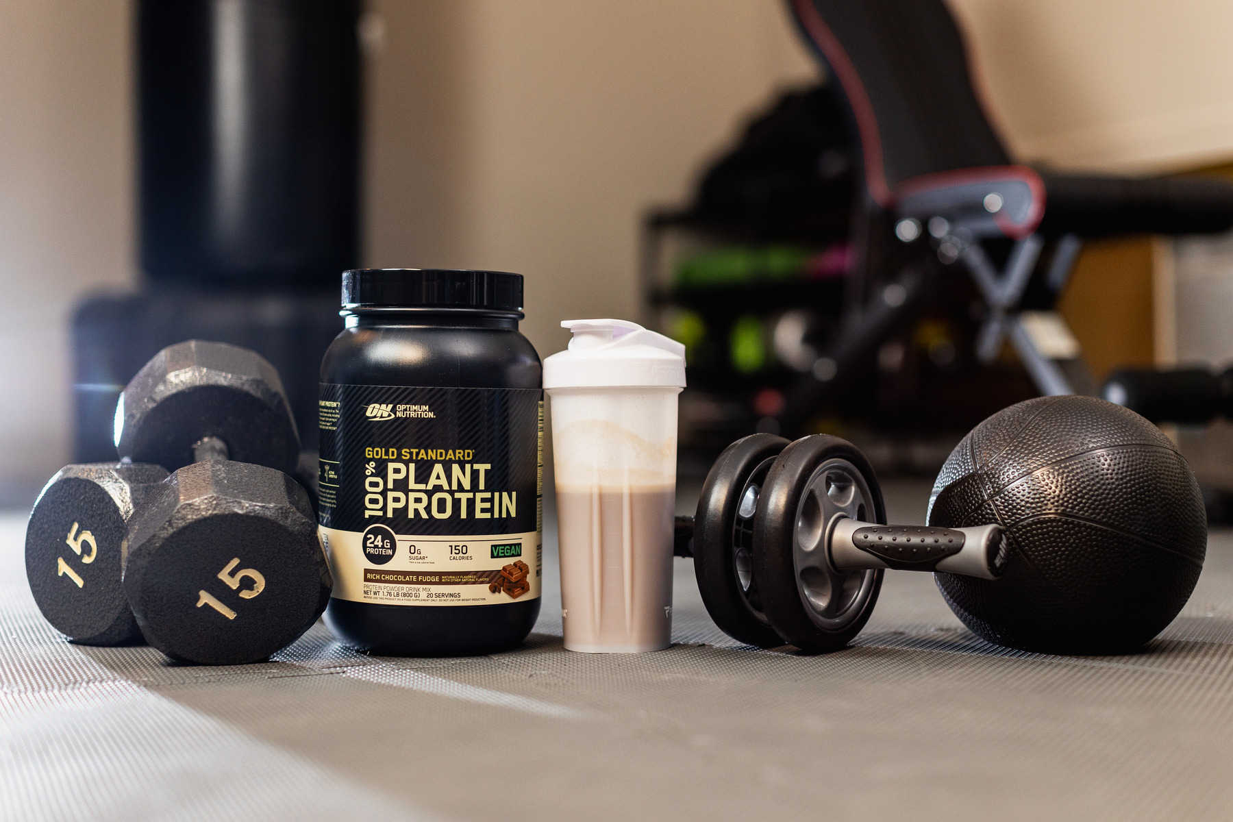Gold Standard Plant Protein Powder container next to a protein shake, weights, and ab roller on a gym floor