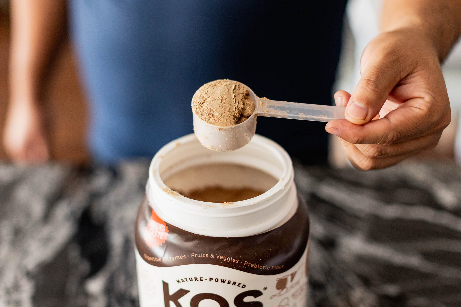 A person's hand holding a scoop of KOS Organic Superfood Plant Protein powder above the open container, with a marbled countertop in the background