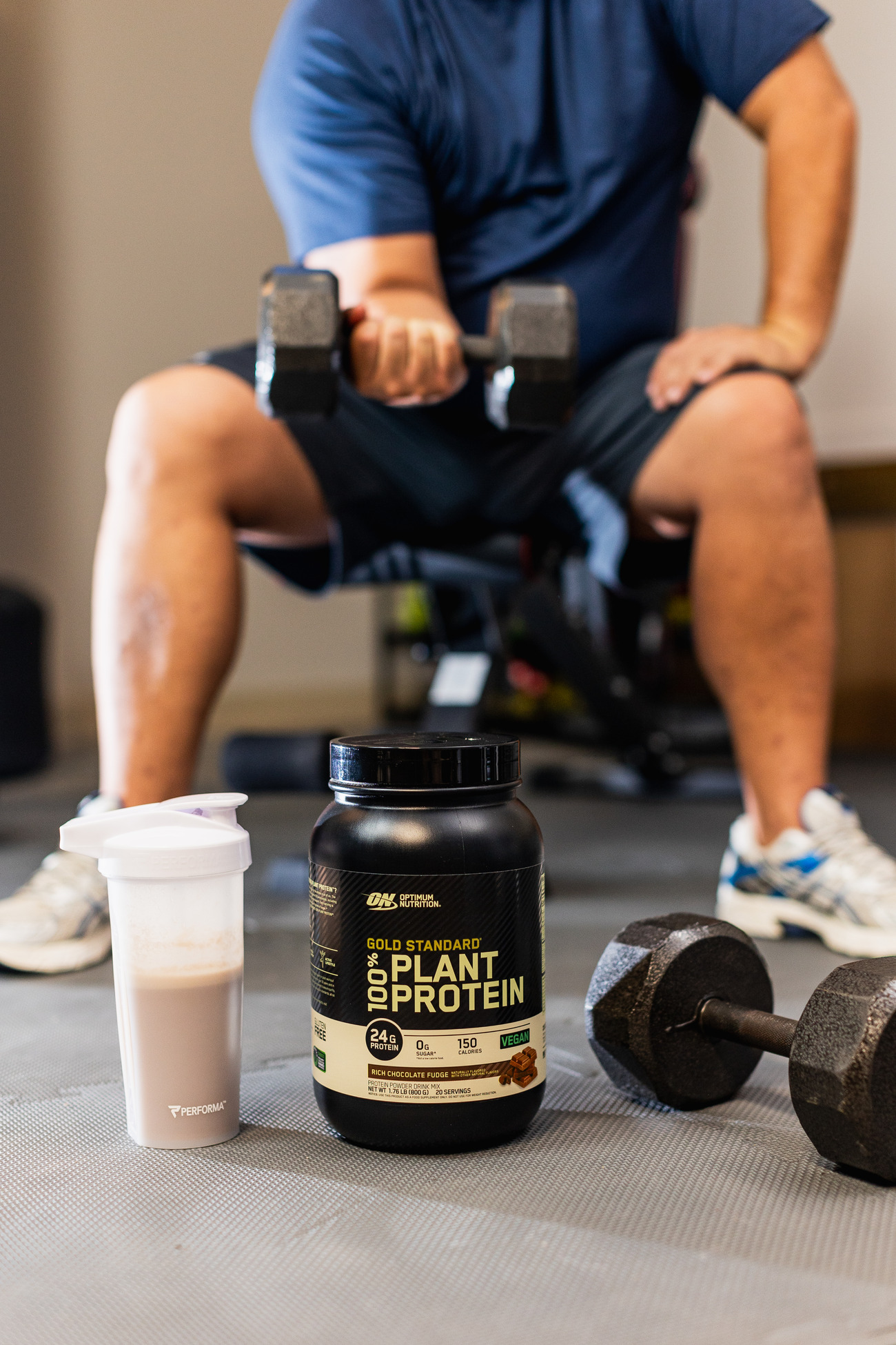 A gym setting with a person seated holding a pair of dumbbells, with a white protein shaker and a black container of Optimum Nutrition Gold Standard Plant Protein in the foreground