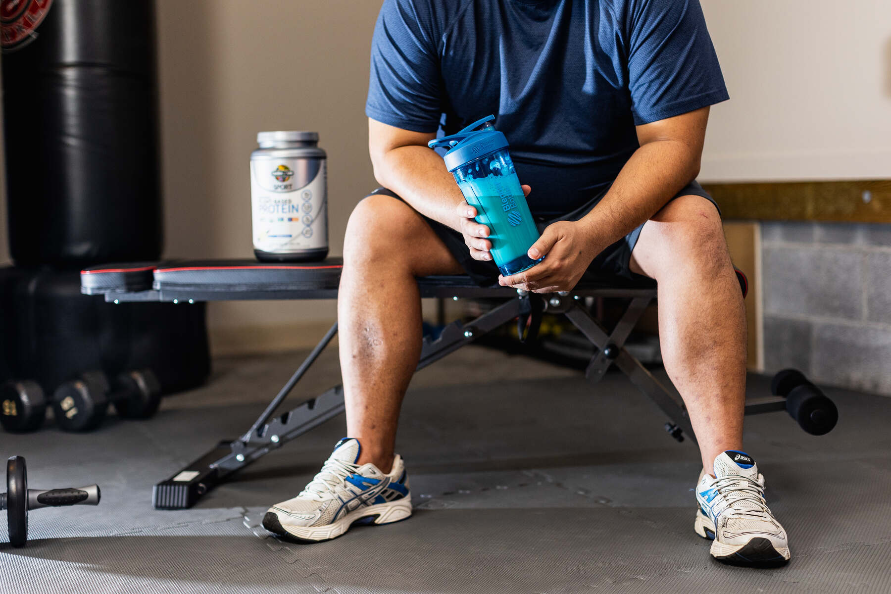 A person in a blue shirt seated in a gym holding a blue protein shaker with a silver container of Garden of Life Sport Organic Plant-Based Protein and workout equipment in the background