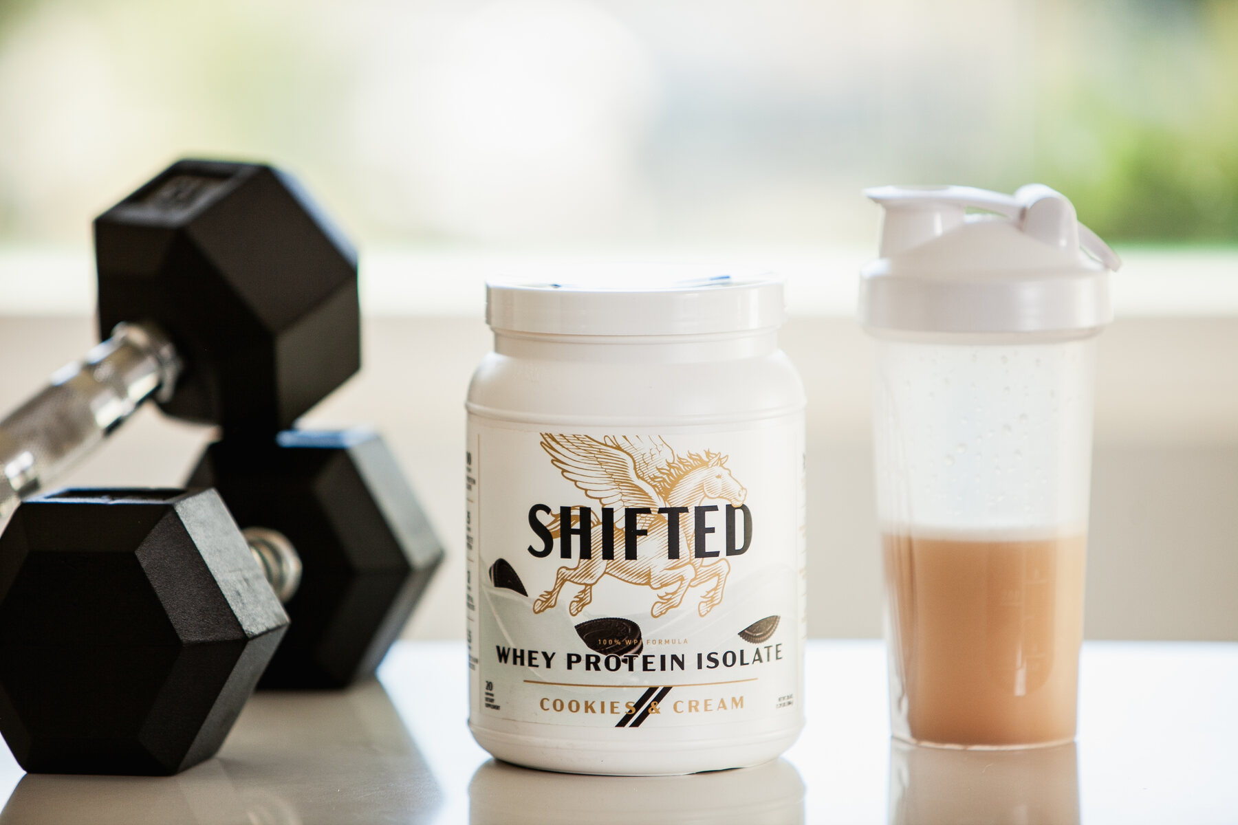 A container of Shifted whey protein isolate next to a pair of dumbbells and a bottle blender filled with a protein drink
